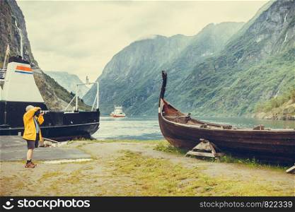 Male tourist with camera taking photo of old wooden viking boat on seashore in norwegian nature, foggy misty day. Mountains and fjord Sognefjord. Tourism and traveling concept. Man taking photo from old viking boat in norway