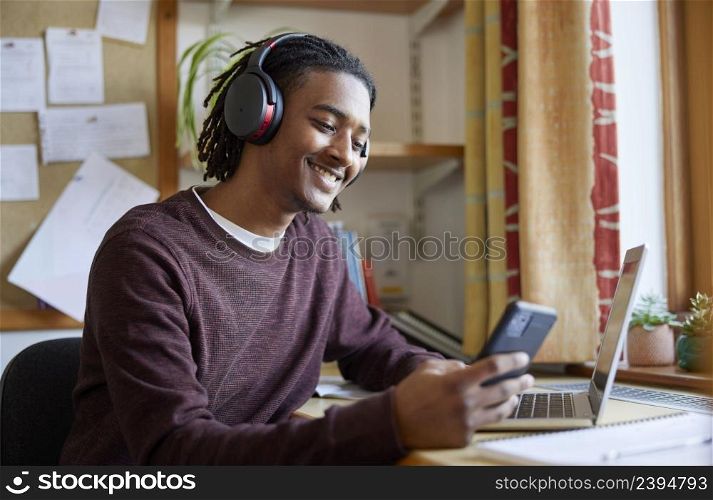 Male University Or College Student Wearing Wireless Headphones With Laptop At Desk In Room Looking At Mobile Phone