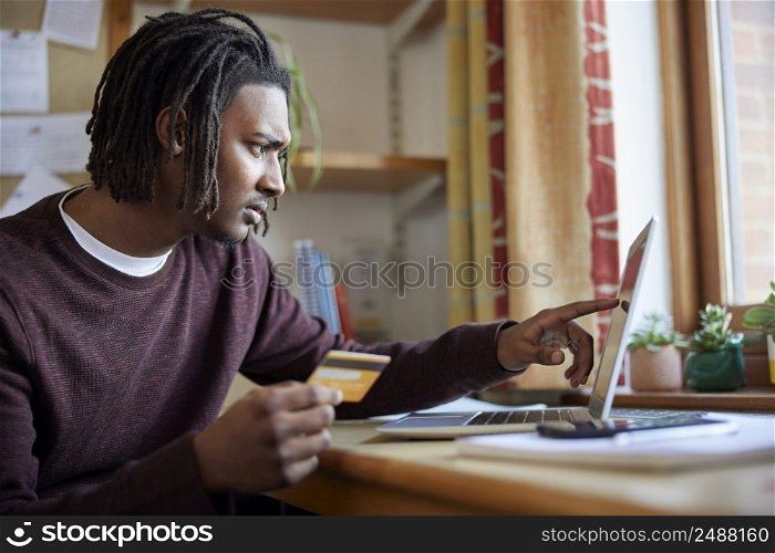 Male University Or College Student With Credit Card Looking At Laptop Worried About Debt At Desk In Room
