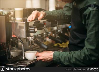 Male waiter standing against coffee machine and cooking a coffee.