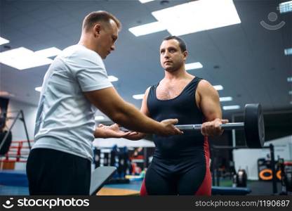 Male weightlifter lies on bench, exercise with dumbbell under instructor control, gym interior on background. Weightlifting workout in sport or fitness club. Weightlifter lies on bench, exercise with dumbbell