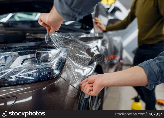 Male worker applies car protection film on front fender. Installation of coating that protects the paint of automobile from scratches. New vehicle in garage, tuning procedure. Worker applies car protection film on front fender