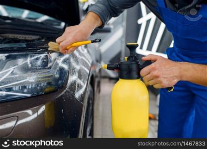 Male worker cleans car surface with spray and brush, preparation before applying of protection film. Installation of coating that protects the paint of automobile from scratches. Vehicle in garage. Worker cleans car surface with spray and brush