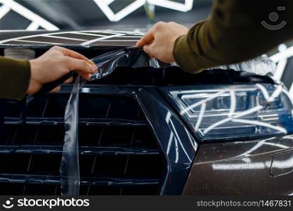 Male worker cuts transparent protection film on car hood. Installation of coating that protects the paint of automobile from scratches. New vehicle in garage, tuning procedure. Worker cuts protection film on car hood
