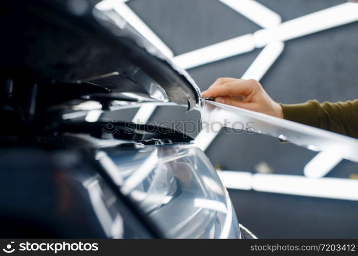 Male worker cuts transparent protection film on car hood. Installation of coating that protects the paint of automobile from scratches. New vehicle in garage, autotuning. Worker cuts protection film on car hood