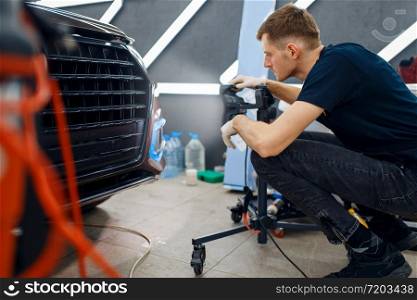 Male worker dries car protection film using a powerful lamp. Installation of coating that protects the paint of automobile from scratches. Vehicle in garage, auto tuning procedure. Male worker dries car protection film, auto tuning