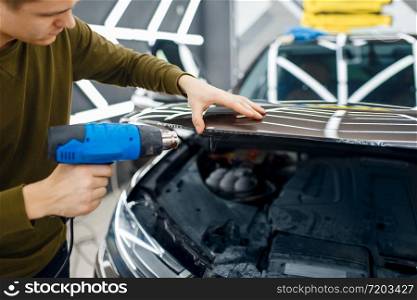 Male worker dries vinyl car protection film on hood. Installation of coating that protects the paint of automobile from scratches. New vehicle in garage, tuning procedure. Male worker dries car protection film on hood