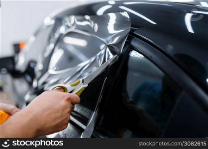 Male worker holds sheet of film, car tinting installation, tuning service. Mechanic applying vinyl tint on vehicle window in garage, tinted automobile glass. Worker holds film, car tinting installation