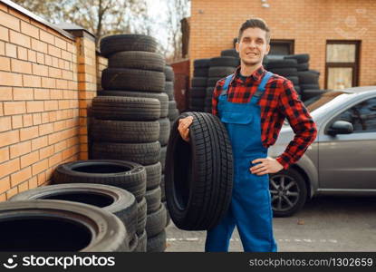 Male worker in uniform at the stack of tyres, tire service. Vehicle repair service or business, man repairing broken wheel. Male worker at the stack of tyres, tire service