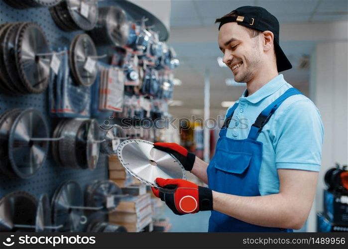 Male worker in uniform choosing edged disc for saw in tool store. Choice of professional equipment in hardware shop, instrument supermarket. Male worker choosing disc for saw in tool store