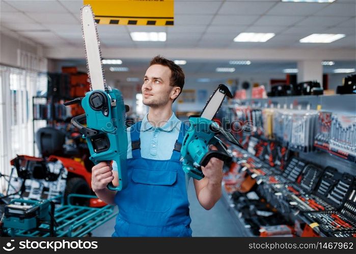 Male worker in uniform holds big and small chainsaws in tool store. Choice of professional equipment in hardware shop, instrument supermarket. Worker holds big and small chainsaws in tool store