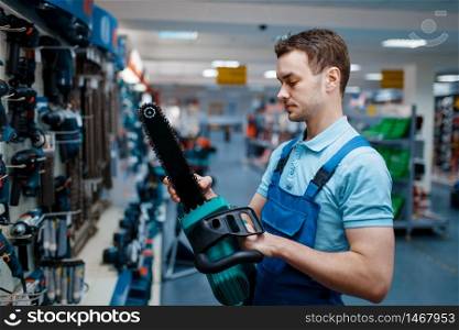 Male worker in uniform holds electric saw in tool store. Choice of professional equipment in hardware shop, instrument supermarket. Worker in uniform holds electric saw in tool store