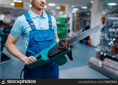 Male worker in uniform holds electric saw in tool store. Choice of professional equipment in hardware shop, instrument supermarket. Worker in uniform holds electric saw in tool store