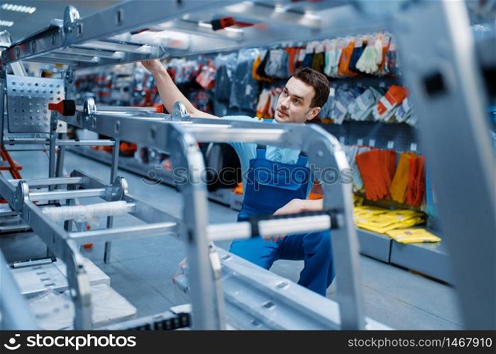 Male worker in uniform holds new aluminum stepladders in tool store. Department with ladders, choice of equipment in hardware shop, instrument supermarket. Worker holds aluminum stepladders in tool store