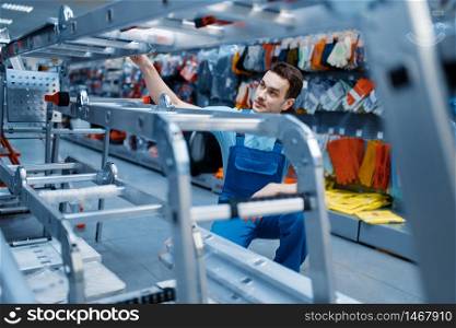 Male worker in uniform holds new aluminum stepladders in tool store. Department with ladders, choice of equipment in hardware shop, instrument supermarket. Worker holds aluminum stepladders in tool store