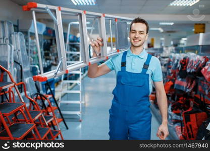 Male worker in uniform holds new aluminum stepladders in tool store. Department with ladders, choice of equipment in hardware shop, instrument supermarket. Worker holds aluminum stepladders in tool store