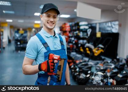 Male worker in uniform holds pneumatic nailer in tool store. Choice of professional equipment in hardware shop, instrument supermarket. Male worker holds pneumatic nailer in tool store