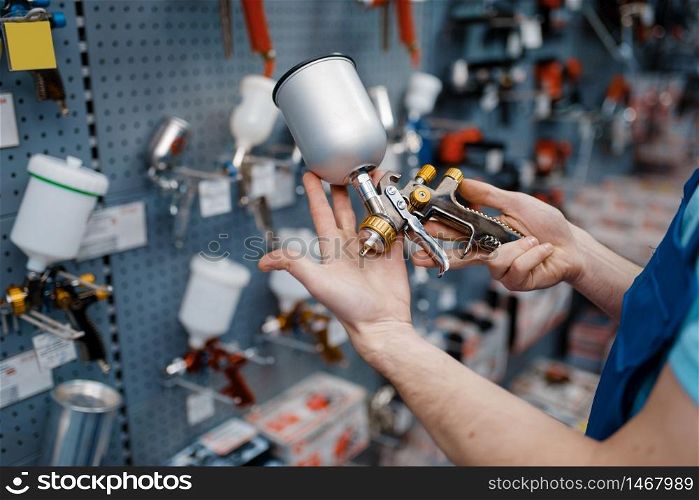 Male worker in uniform holds pneumatic paint spray gun in tool store. Choice of professional equipment in hardware shop, instrument supermarket. Male worker holds pneumatic paint gun, tool store
