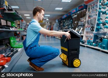 Male worker in uniform holds toolbox on wheels in tool store. Choice of professional equipment in hardware shop, instrument supermarket. Male worker holds toolbox on wheels in tool store