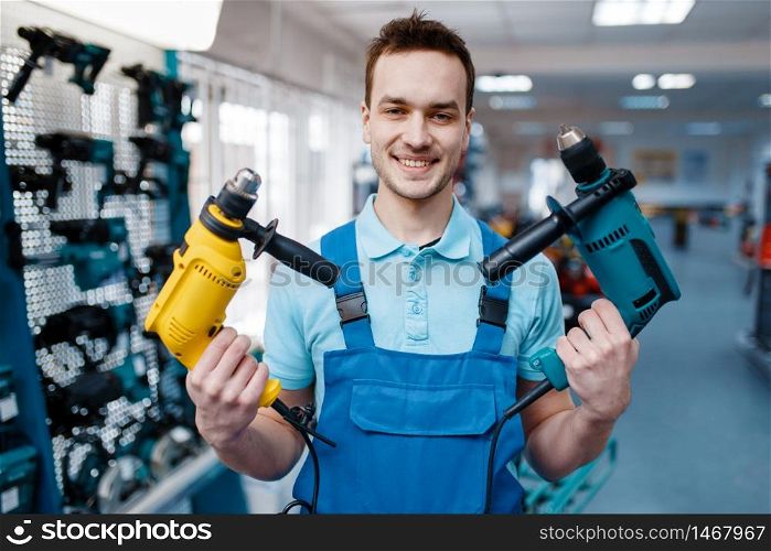 Male worker in uniform holds two electric drills in tool store. Choice of professional equipment in hardware shop, instrument supermarket. Worker holds two electric drills in tool store