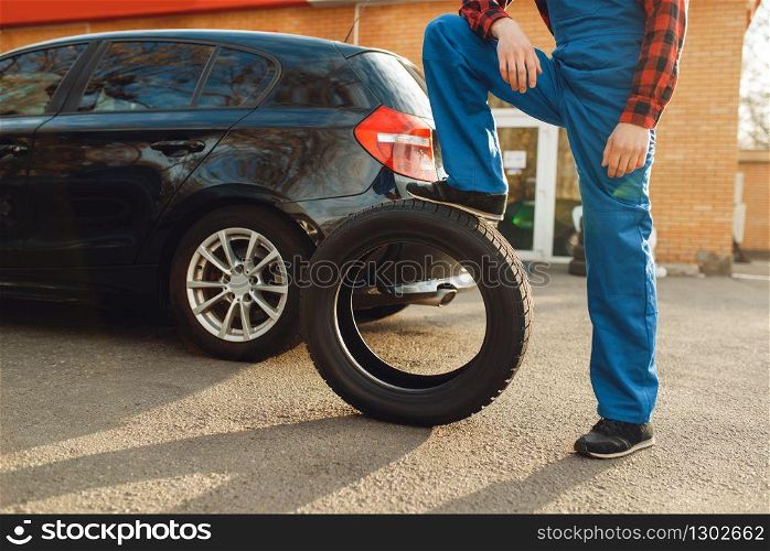 Male worker in uniform poses with tyre, tire service. Vehicle repair service or business, man change wheels on automobile outdoor. Worker in uniform poses with tyre, tire service