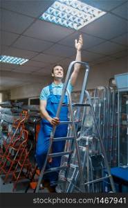 Male worker in uniform standing on stepladder in tool store. Department with ladders, choice of equipment in hardware shop, instrument supermarket. Male worker standing on stepladder in tool store