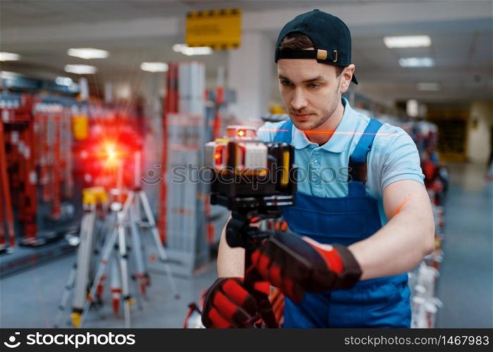 Male worker in uniform testing laser level on tripod in tool store. Choice of professional equipment in hardware shop, instrument supermarket. Worker testing laser level on tripod in tool store