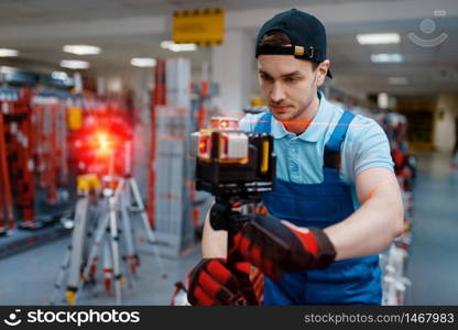 Male worker in uniform testing laser level on tripod in tool store. Choice of professional equipment in hardware shop, instrument supermarket. Worker testing laser level on tripod in tool store