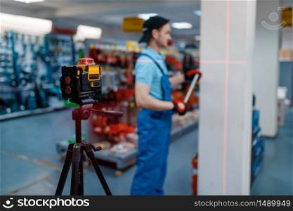 Male worker in uniform testing laser level on tripod in tool store. Choice of professional equipment in hardware shop, instrument supermarket. Worker testing laser level on tripod in tool store