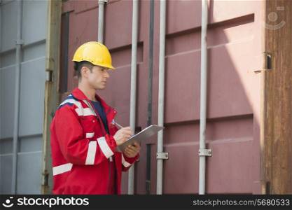 Male worker inspecting cargo container while writing on clipboard in shipping yard