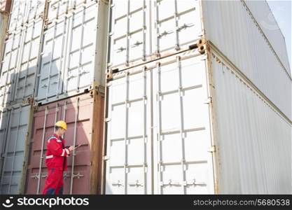Male worker inspecting cargo containers while writing on clipboard in shipping yard