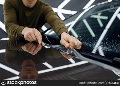 Male worker installs transparent protection film on car hood. Installation of coating that protects the paint of automobile from scratches. New vehicle in garage, tuning procedure. Worker installs protection film on car hood