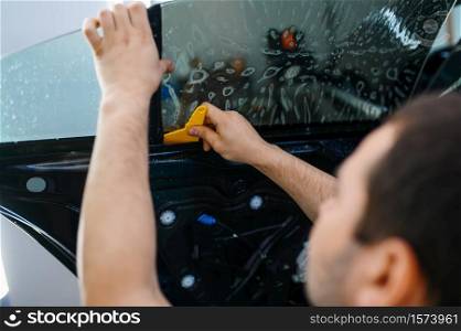 Male worker installs wetted car tinting, tuning service. Mechanic applying vinyl tint on vehicle window in garage, tinted glass. Worker installs wetted car tinting, tuning service