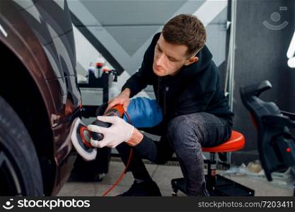 Male worker polishes bumper using polishing machine, car detailing. Preparationa before installation of coating that protects the paint of automobile from scratches. Vehicle in garage, auto tuning. Male worker polishes bumper, car detailing