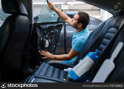 Male worker prepares car for tinting, tuning service. Mechanic applying vinyl tint on vehicle window in garage, tinted automobile glass. Worker prepares car for tinting, tuning service