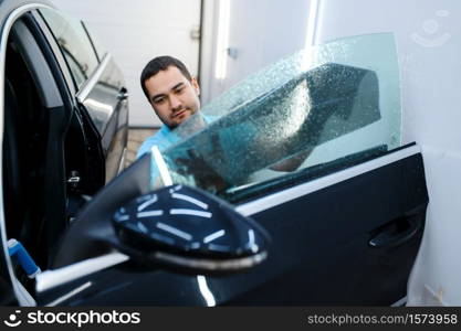 Male worker tries on sheet of car tinting, tuning service. Mechanic applying vinyl tint on vehicle window in garage, tinted automobile glass. Male worker tries on sheet of car tinting