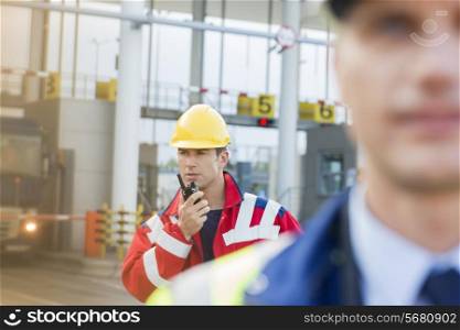 Male worker using walkie-talkie with colleague in foreground at shipyard