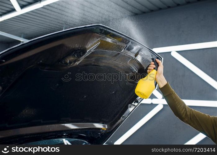 Male worker wets car hood surface with spray before applying of protection film. Installation of coating that protects the paint of automobile from scratches. New vehicle in garage, tuning procedure. Male worker wets car hood surface with spray
