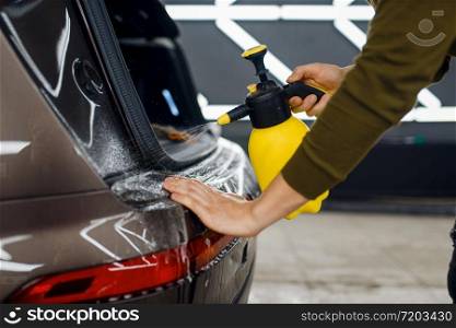 Male worker wets rear bumper surface of car with spray before applying of protection film. Installation of coating that protects the paint of automobile from scratches. Vehicle in garage, detailing. Worker wets rear bumper surface of car, detailing