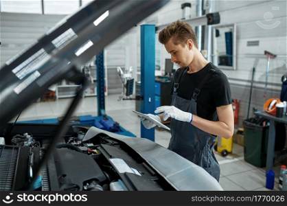 Male worker with checklist stands at the hood, car service. Vehicle repairing garage, man in uniform, automobile station interior on background. Worker with checklist stands at hood, car service