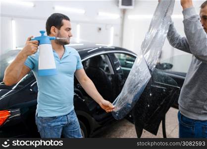 Male worker with spray wetting car tinting, tuning service. Mechanic applying vinyl tint on vehicle window in garage, tinted automobile glass. Male worker with spray wetting car tinting