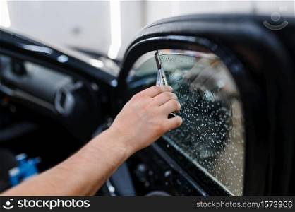 Male worker with squeegee wipes car tinting, tuning service. Mechanic applying vinyl tint on vehicle window in garage, tinted automobile glass. Male worker with squeegee wipes car tinting