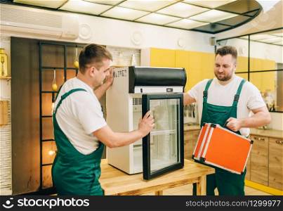 Male workers in uniform repair refrigerator at home. Repairing of fridge occupation, professional service. Male workers in uniform repair refrigerator