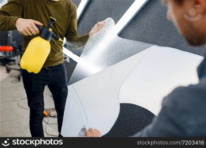 Male workers wets car protection film before applying. Installation of coating that protects the paint of automobile from scratches. New vehicle in garage, tuning procedure. Workers wets car protection film before applying