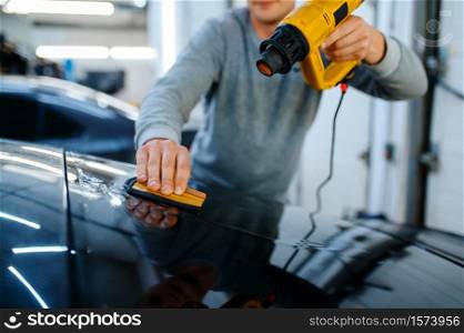 Male wrapper holds squeegee and heat gun, car tinting service. Worker applying vinyl tint on vehicle window in garage, tinted automobile glass. Wrapper holds squeegee and heat gun, car tinting