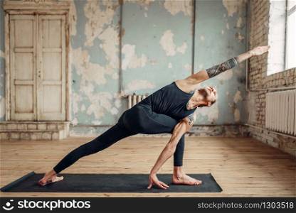 Male yoga doing flexibility exercise on mat in gym with grunge interior. Fit workout indoors, yogi studio