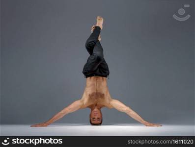 Male yoga standing on his head and hands, meditation, grey background. Strong man doing yogi exercise, asana training, top concentration, healthy lifestyle. Male yoga standing on his head and hands