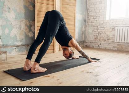 Male yoga with tattoo on hand doing stretching exercise on mat in gym with grunge interior. Fit workout indoors. Male yoga doing stretching exercise on mat