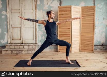 Male yoga with tattoo on hand doing stretching exercise on mat in gym with grunge interior. Fit workout indoors. Male yoga doing stretching exercise on mat