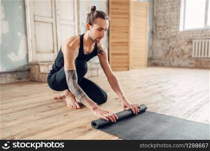 Male yoga with tattoo on hand prepares mat for training in gym with grunge interior. Fit workout indoors. Male yoga with tattoo prepares mat for training